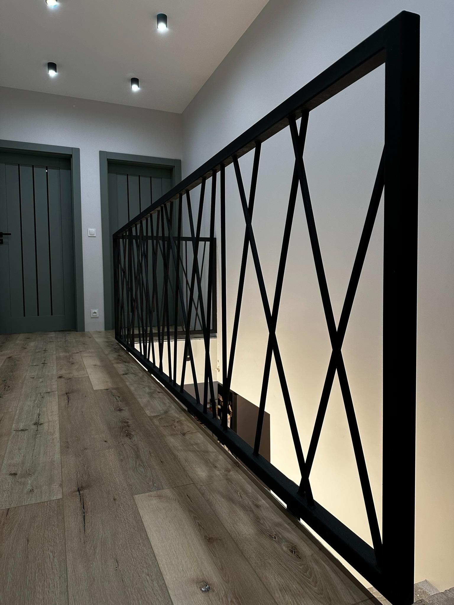 Nowoczesna balustrada schodowa z czarnego metalu o geometrycznym wzorze krzyżujących się prętów, drewniana podłoga, widoczne drzwi w tle.