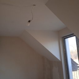 Mariusz Godzina - Poddasze w trakcie wykańczania: surowe ściany, okno z widokiem na zewnątrz, wisząca żarówka na kablu. Widoczny skos dachu.