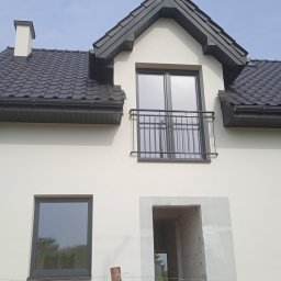 Mariusz Godzina - Nowy dom jednorodzinny z czarną dachówką, oknami i balkonem, w trakcie wykańczania elewacji, widoczne rusztowania i niezakończone prace.