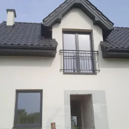 Nowy dom jednorodzinny z czarną dachówką, oknami i balkonem, w trakcie wykańczania elewacji, widoczne rusztowania i niezakończone prace.