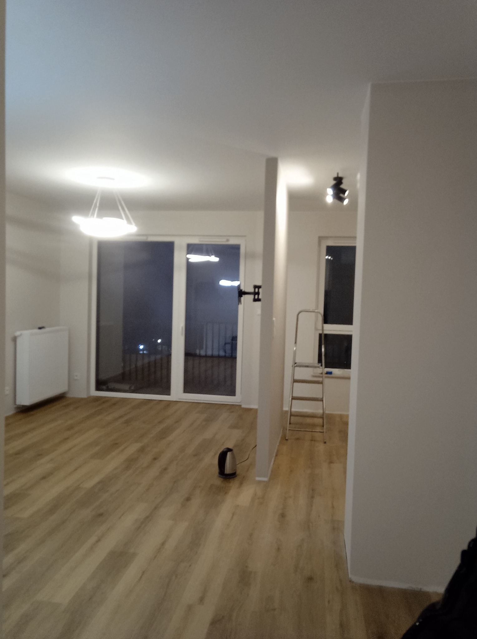 Pomalowane na biało wnętrze mieszkania z panelami podłogowymi, widoczne okno balkonowe z widokiem na miasto nocą, grzejnik, drabina malarska i lampa sufitowa.