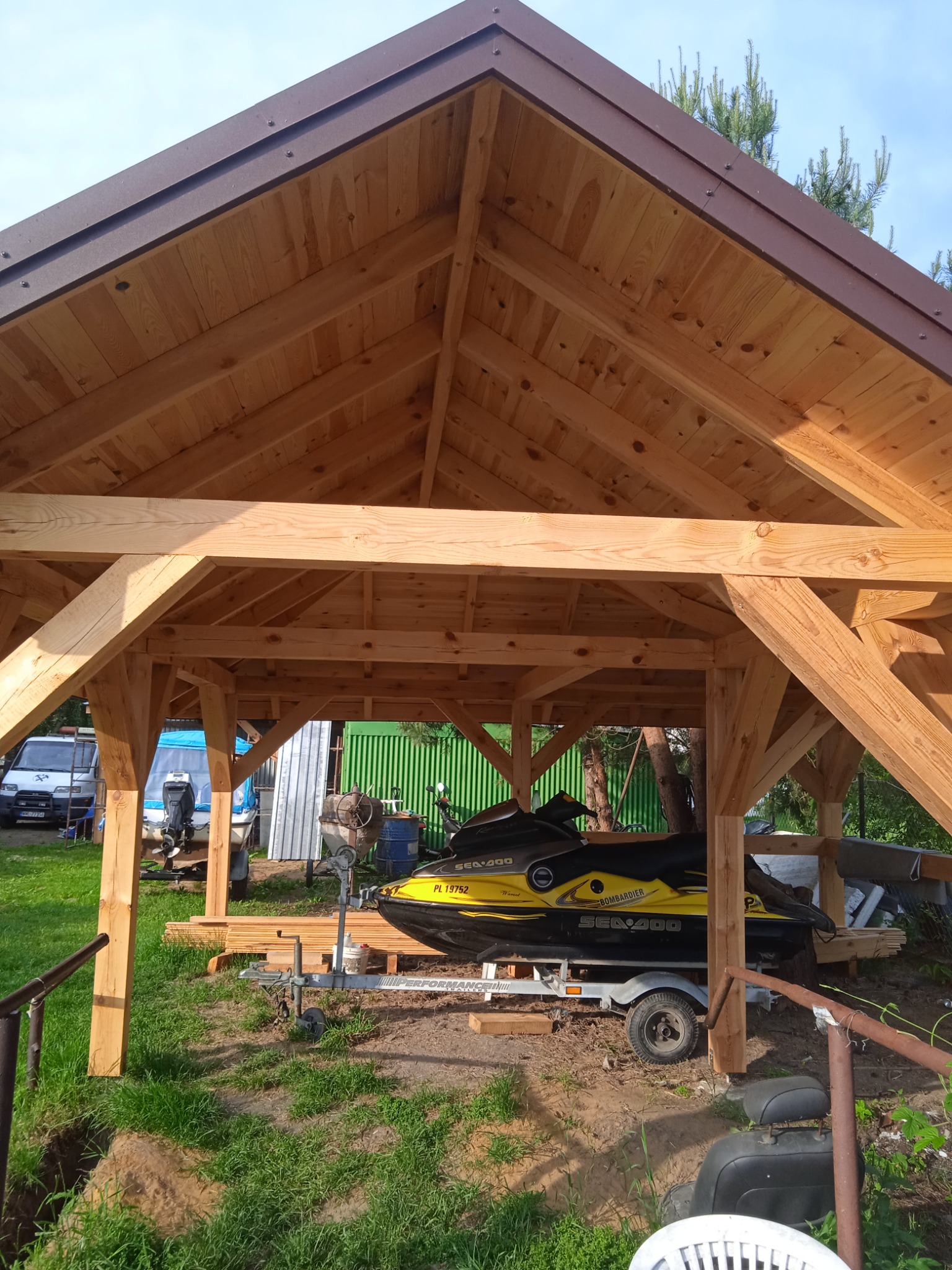 Drewniana konstrukcja z brązowym dachem, prawdopodobnie wiata lub altana, wewnątrz skuter wodny na przyczepie, w tle widoczny samochód dostawczy i inne przedmioty.
