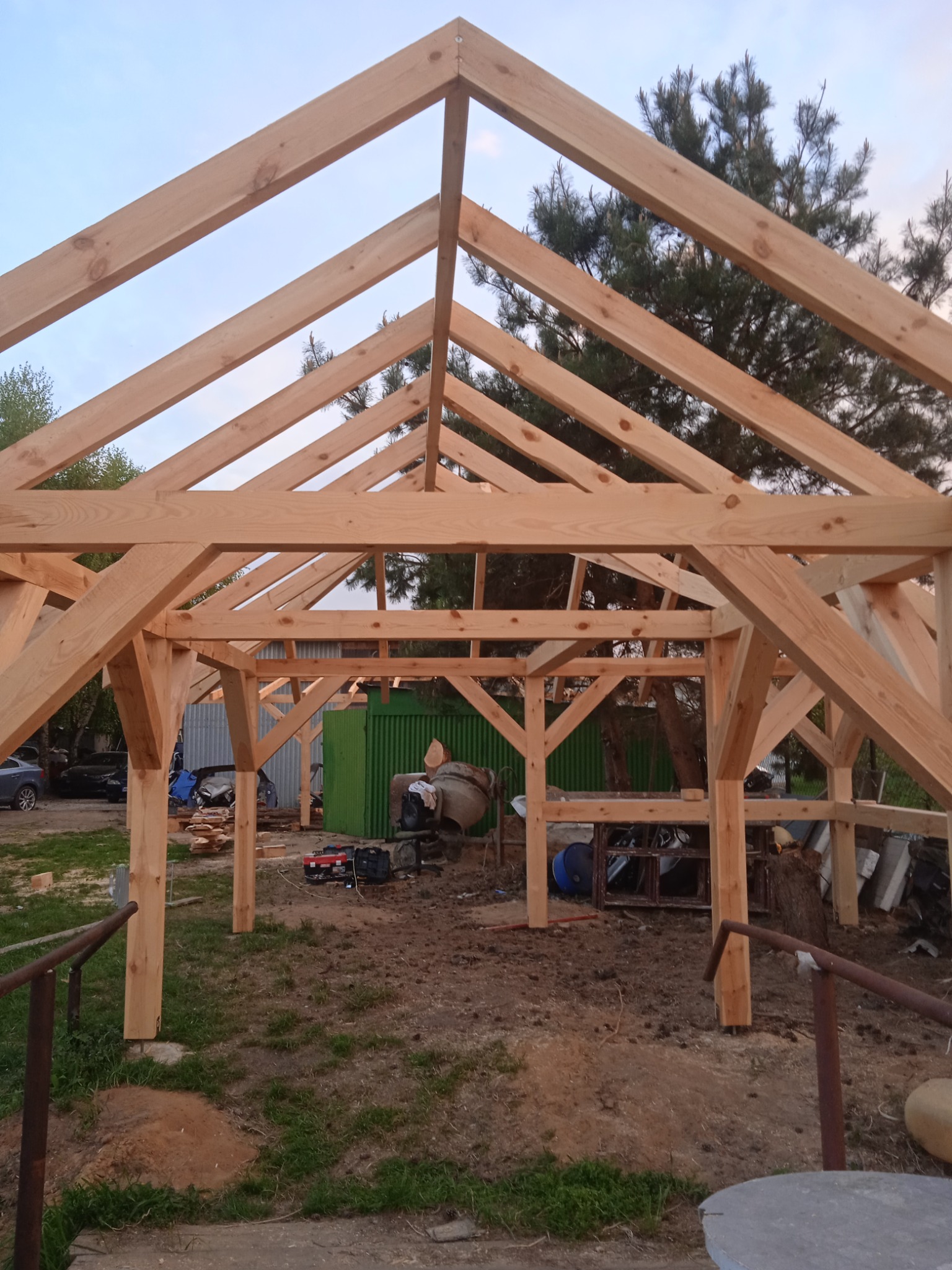 Surowa konstrukcja drewniana dachu w trakcie montażu, widoczne elementy więźby dachowej z jasnego drewna na tle zieleni i błękitnego nieba, na ziemi rozłożone narzędzia i materiały budowlane.