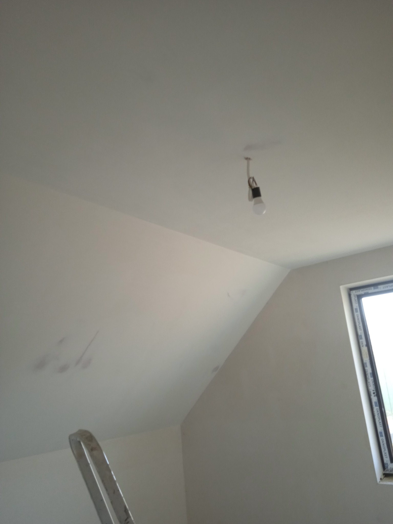 Poddasze z oknem po szpachlowaniu, widoczne niedoskonałości przed malowaniem, drabina w rogu, surowa żarówka