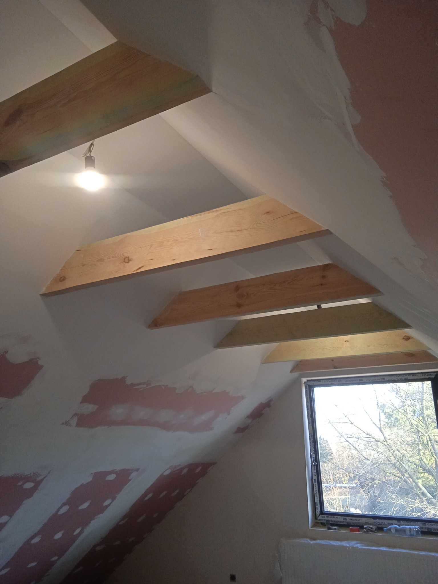Poddasze w trakcie wykańczania: widoczne drewniane elementy konstrukcyjne dachu, ściany przygotowane do malowania z widocznymi warstwami szpachli, okno z widokiem na drzewa.