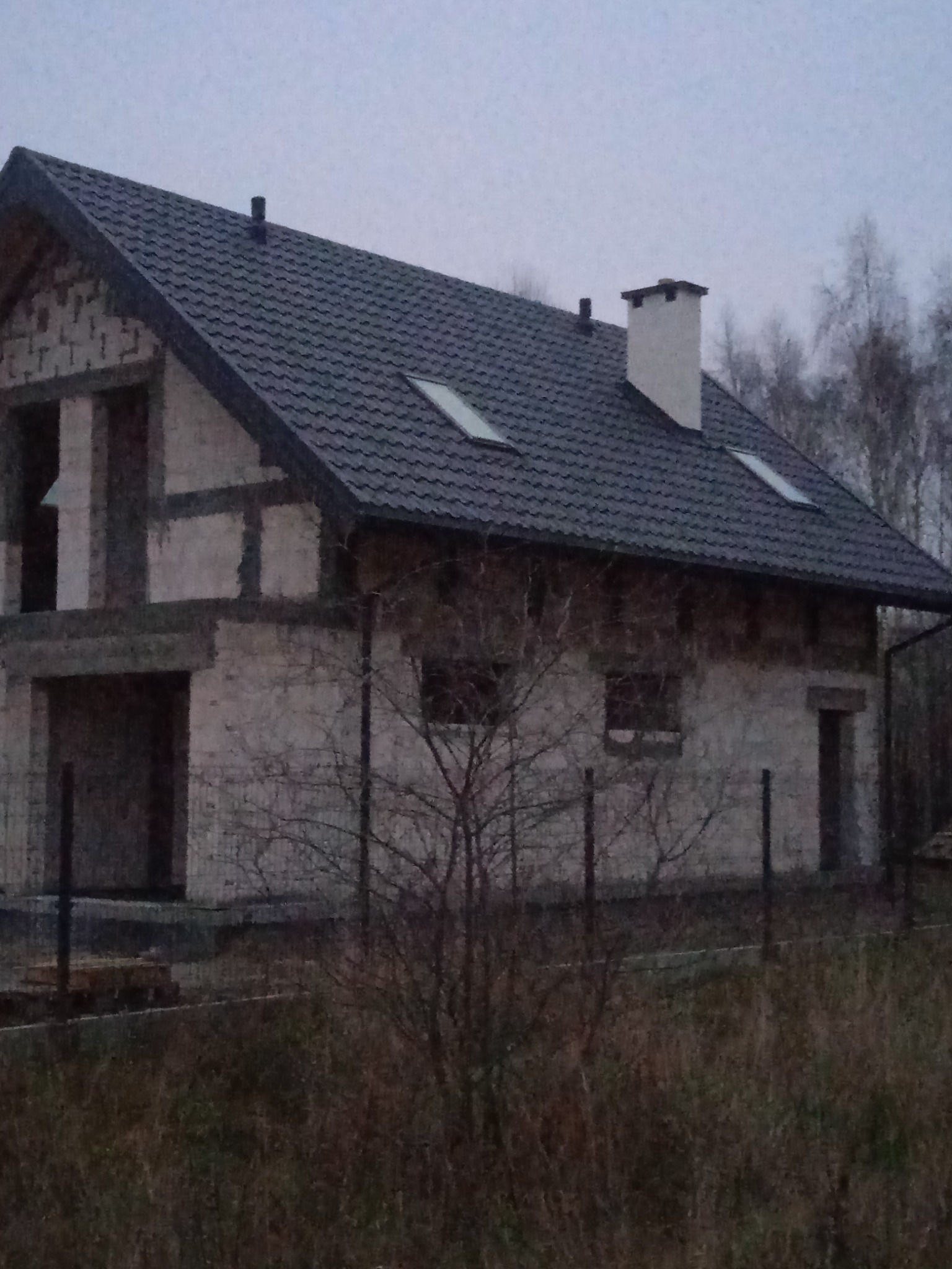 Surowy stan budynku jednorodzinnego z ciemną dachówką i kominem, widoczne okna dachowe, ściany z pustaków, brak wykończenia elewacji, jesienne otoczenie z drzewami bez liści, pochmurne niebo.