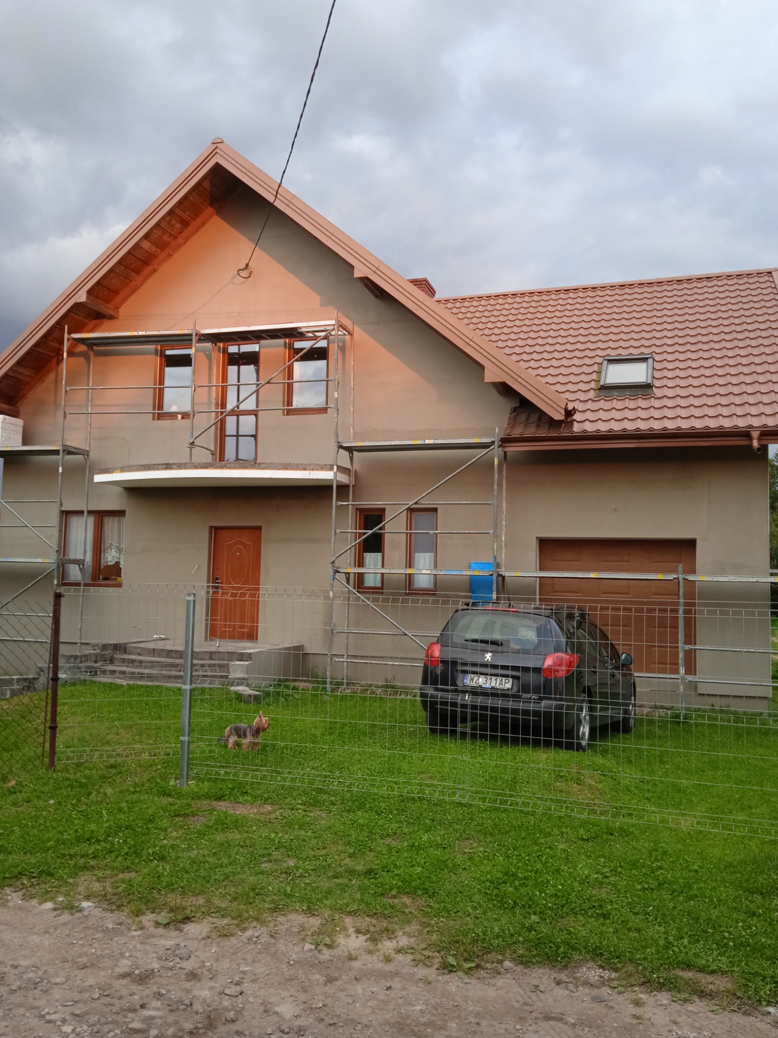 Dom jednorodzinny w trakcie ocieplania elewacji, widoczne rusztowania, częściowo otynkowana ściana, brązowy dach, zaparkowany samochód i pies rasy Yorkshire Terrier na trawniku.