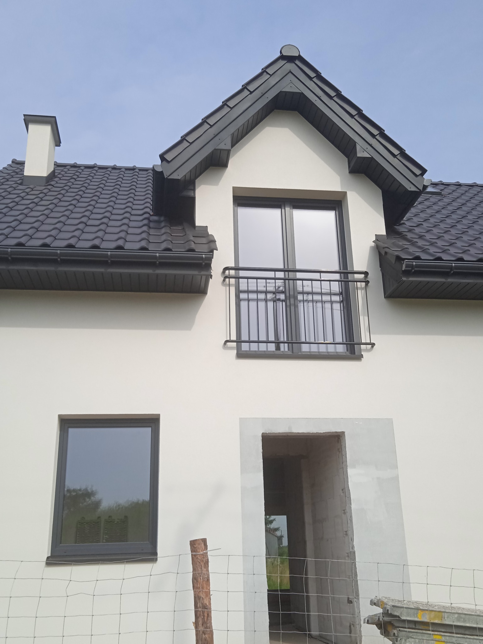 Nowy dom jednorodzinny z czarną dachówką, oknami i balkonem, w trakcie wykańczania elewacji, widoczne rusztowania i niezakończone prace.