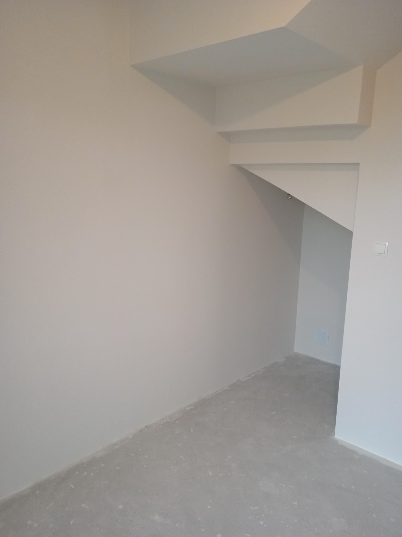 Pomalowane na biało wnętrze ze świeżo wykończoną podłogą, widoczne załamania ścian i sufitu oraz wnęka z oświetleniem punktowym.