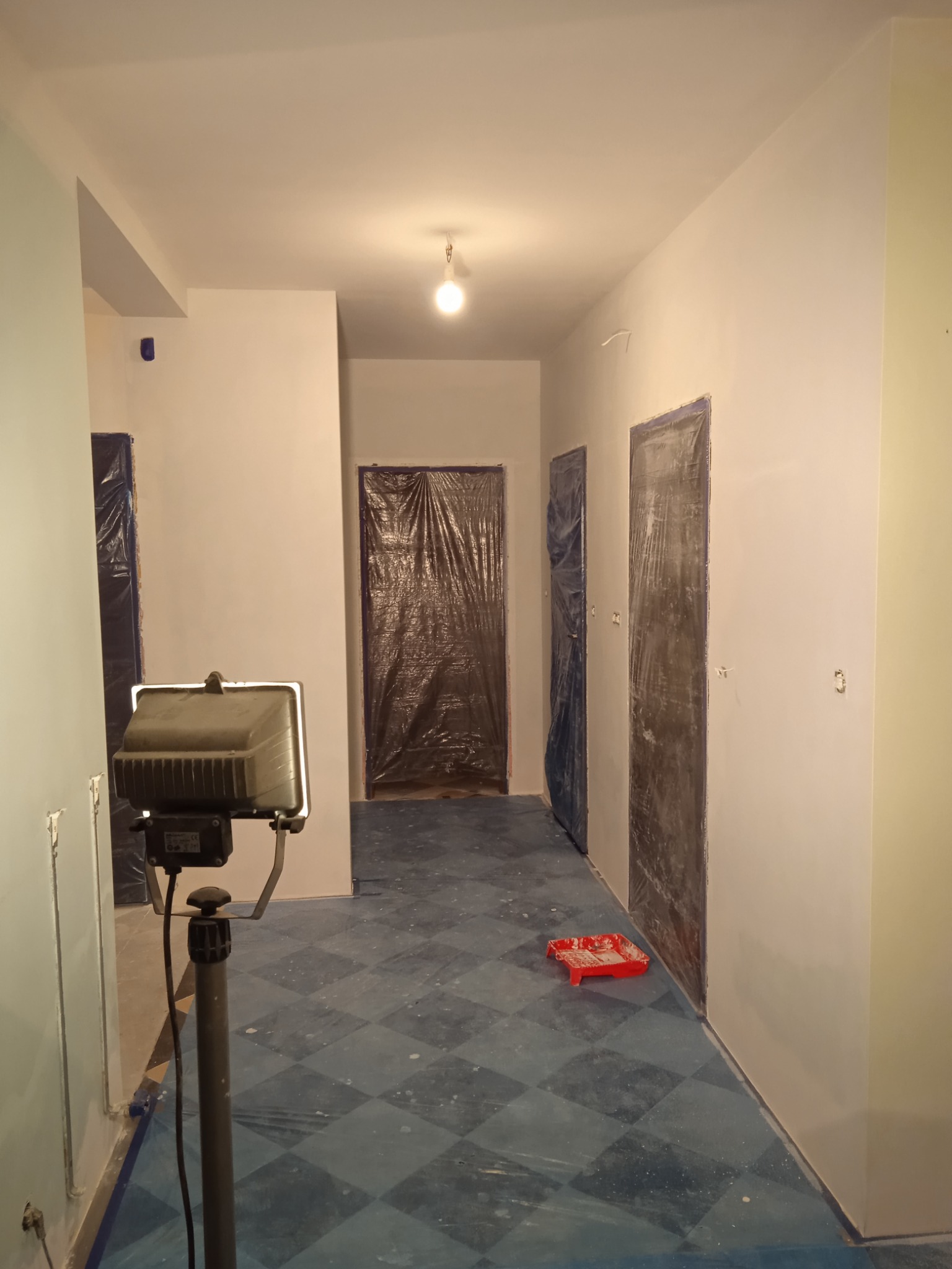 Korytarz w trakcie odświeżania, ściany pomalowane na jasny kolor, drzwi zabezpieczone folią, podłoga z wzorem w romby, widoczny halogen na statywie i kuweta malarska.