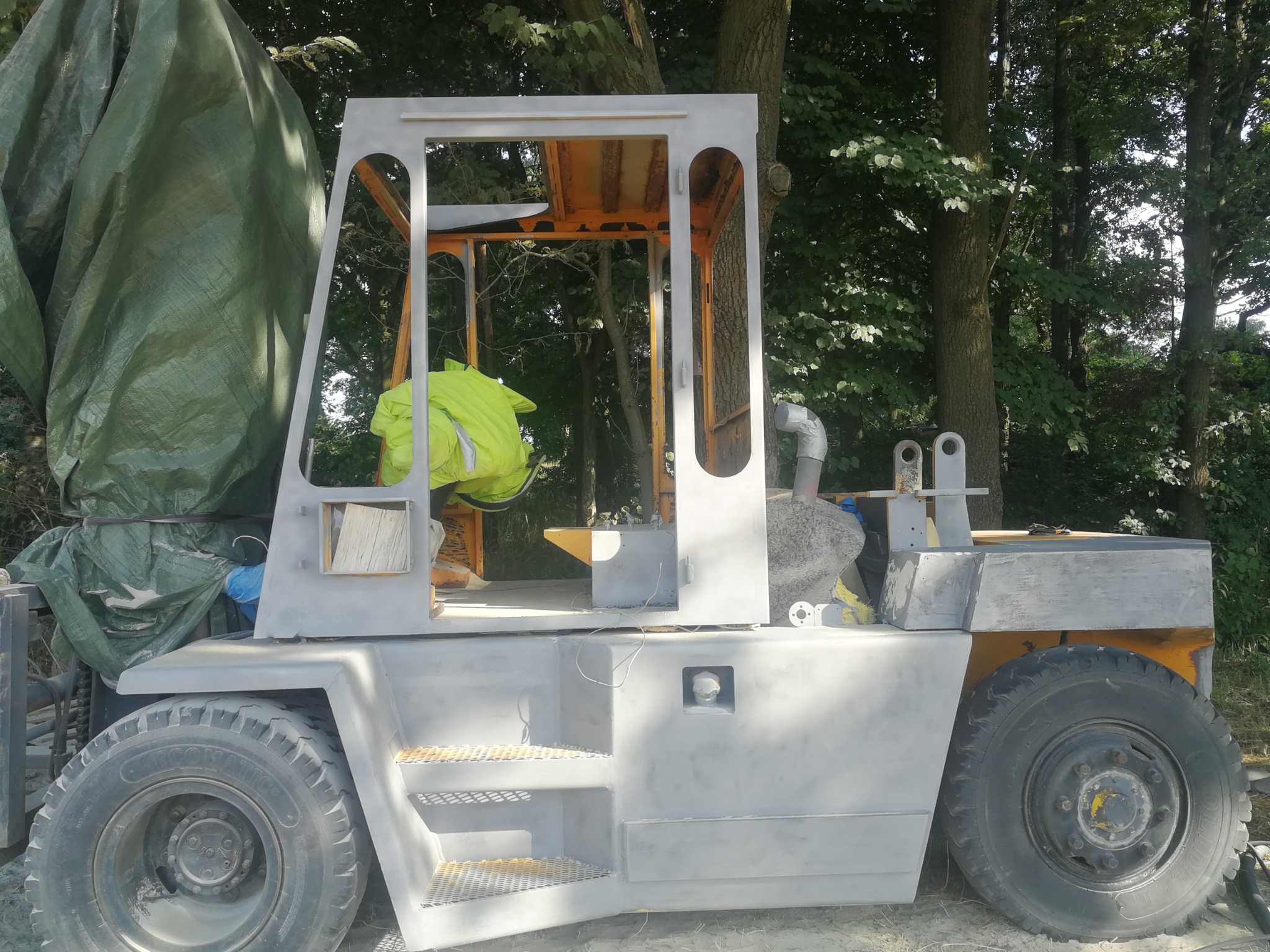 Wózek widłowy w trakcie przygotowania do malowania, zabezpieczony folią, po nałożeniu podkładu, widoczna osoba w odblaskowej kamizelce w kabinie.