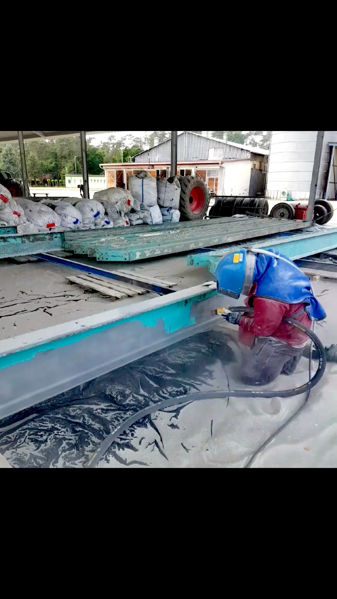 Pracownik w kombinezonie ochronnym i hełmie obrabia strumieniem ściernym metalową konstrukcję, widoczne ślady usuniętej farby i nagromadzony piasek.