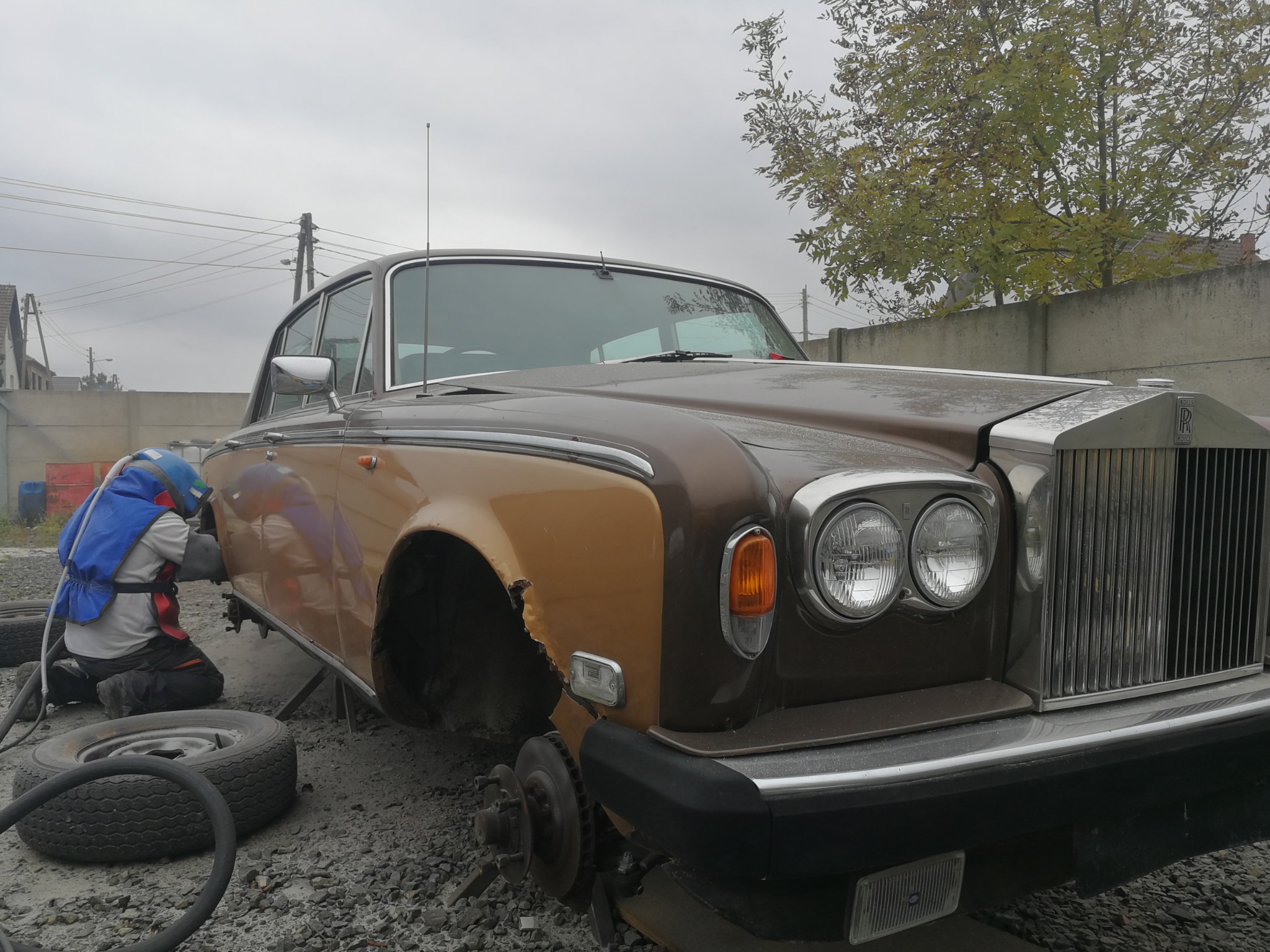 Piaskowanie karoserii brązowo-złotego, zabytkowego Rolls Royce'a na zewnątrz, widoczne uszkodzenia i brak koła.