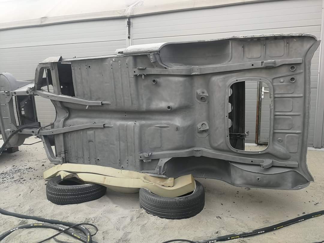 Podwozie samochodu po obróbce, przygotowane do dalszych prac, wsparte na oponach i piankowych podkładkach, widoczne przewody na piaszczystym podłożu.