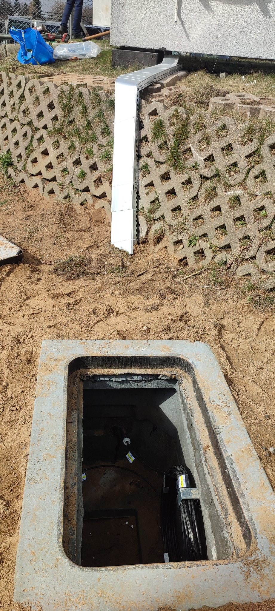 Otwarty betonowy właz do studzienki kablowej z widocznymi kablami wewnątrz, obok ściana z betonowych ażurów i metalowa rura osłonowa na kable.