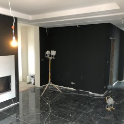 Mdkremonty - Pokój w trakcie odświeżania, pomalowane na czarno ściany, marmurowa podłoga, oświetlenie budowlane, widoczne puszki z farbą i kable.