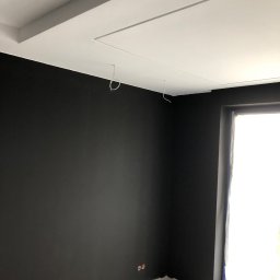 Mdkremonty - Świeżo pomalowane na czarno ściany pokoju z białym sufitem i wystającymi przewodami elektrycznymi, okno zabezpieczone folią malarską, widoczne gniazdka elektryczne.