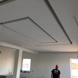 Mdkremonty - Sufit podwieszany z płyt kartonowo-gipsowych z dekoracyjnymi wnękami, widok z dołu w trakcie prac wykończeniowych w pomieszczeniu w Warszawie, z widocznymi kablami i pracownikiem w tle.