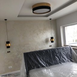 Mdkremonty - Pokój w trakcie prac wykończeniowych: ściana z efektem dekoracyjnym, wiszące lampy z czarnymi i złotymi elementami, zakryte folią łóżko i sufit z podwieszanym elementem.