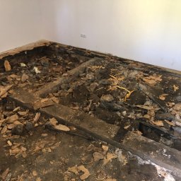 Mdkremonty - Pokój w trakcie demontażu podłogi, widoczne legary, gruz i resztki starej podłogi, ściany w kolorze białym, gniazdko elektryczne na ścianie.