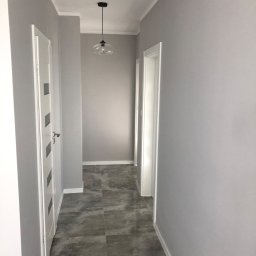 Mdkremonty - Korytarz po remoncie z szarymi ścianami, ciemnoszarą podłogą z płytek imitujących beton i białymi drzwiami z przeszkleniami, oświetlony wiszącą lampą z przezroczystego szkła.