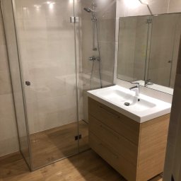 Mdkremonty - Nowoczesna kabina prysznicowa ze szklanymi drzwiami i drewnopodobną podłogą, umywalka nablatowa na szafce z trzema szufladami, lustro bez ramy, kremowe płytki na ścianach.