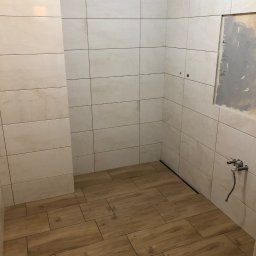 Mdkremonty - Wnętrze łazienki w trakcie prac wykończeniowych: ułożone beżowe płytki imitujące drewno na podłodze, jasne, marmurkowe płytki na ścianach, widoczne podejście wodne i fragment ściany przygotowany...