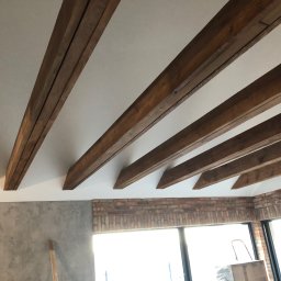 Mdkremonty - Jasne wnętrze z drewnianymi belkami stropowymi i fragmentem ściany z cegły. Widoczna ściana z szarą fakturą. Duże okno w tle.