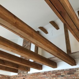 Mdkremonty - Drewniane belki stropowe, widoczne od dołu, w jasnym wnętrzu z fragmentem ściany z cegły. Konstrukcja w trakcie montażu, widoczne przewody.