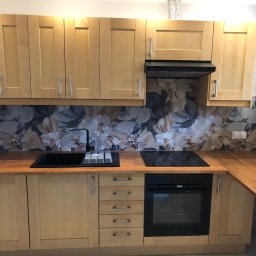 Mdkremonty - Jasna kuchnia z drewnianymi frontami, czarnym zlewem i płytą. Nad blatem dekoracyjne płytki z motywem kwiatowym. Nowoczesny design.