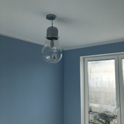 Mdkremonty - Świeżo pomalowane na niebiesko ściany w pokoju z ozdobną lampą w kształcie żarówki, widoczna drabina malarska w rogu przy oknie.