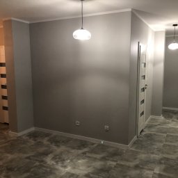 Mdkremonty - Szare wnętrze mieszkania po odświeżeniu, widoczne gładkie ściany, szara podłoga imitująca kamień i białe listwy przypodłogowe oraz sufitowe, oświetlenie stanowią dwie wiszące lampy z mlecznymi...