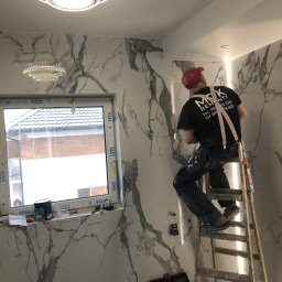 Mdkremonty - Pracownik w czerwonej czapce na drabinie, montuje płytki imitujące marmur w łazience z oknem, widoczne narzędzia i oświetlenie LED.