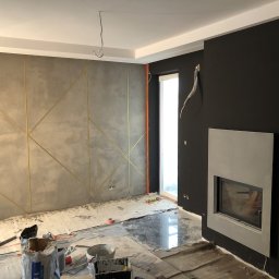 Mdkremonty - Szara ściana z geometrycznym wzorem ze złotych listew w trakcie prac wykończeniowych, widoczne narzędzia i materiały budowlane, czarna ściana z kominkiem
