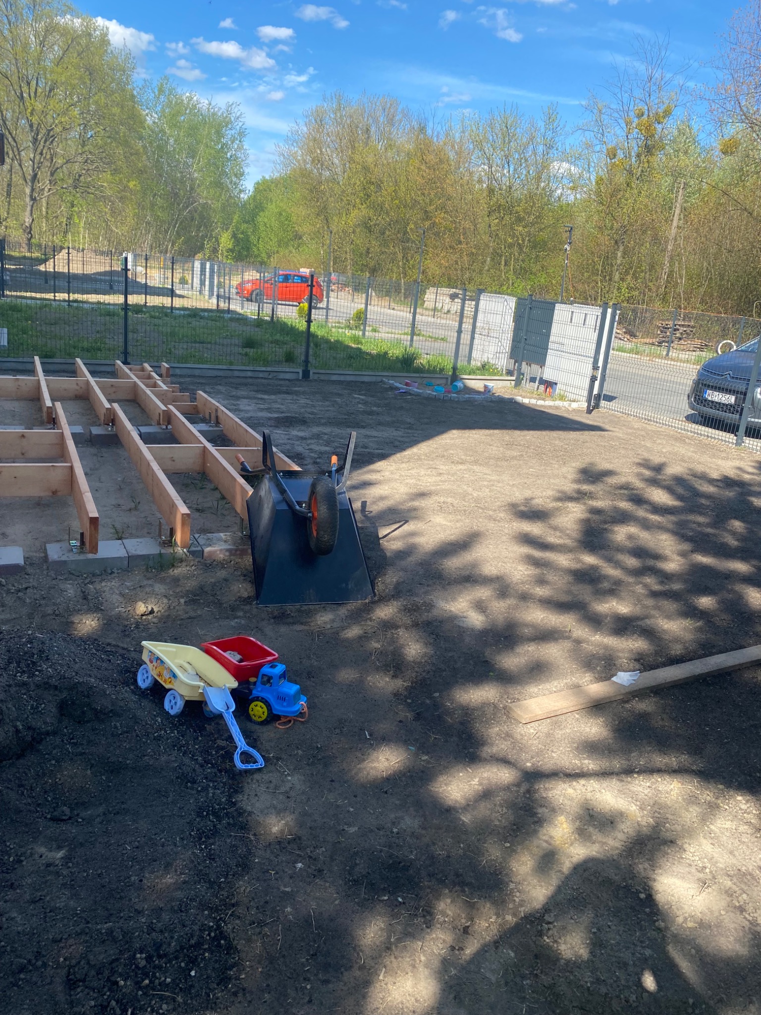 Przygotowanie podłoża pod taras: drewniana konstrukcja ramy na betonowych bloczkach, taczka i zabawki na ziemi, ogrodzenie w tle.