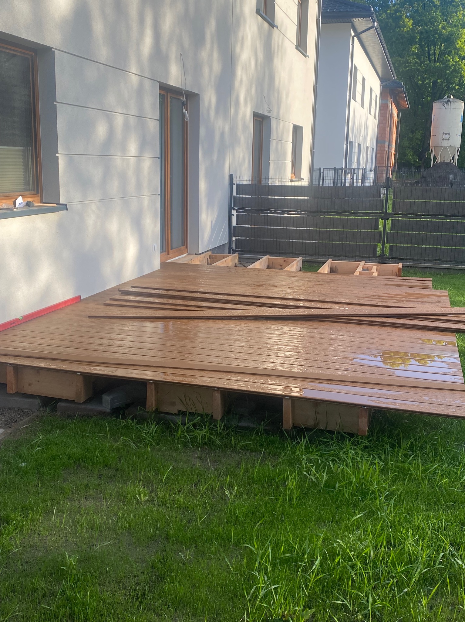 Montaż drewnianego tarasu przy domu jednorodzinnym. Widoczne deski, konstrukcja nośna i poziomica. Częściowo ukończony etap budowy tarasu na trawie.