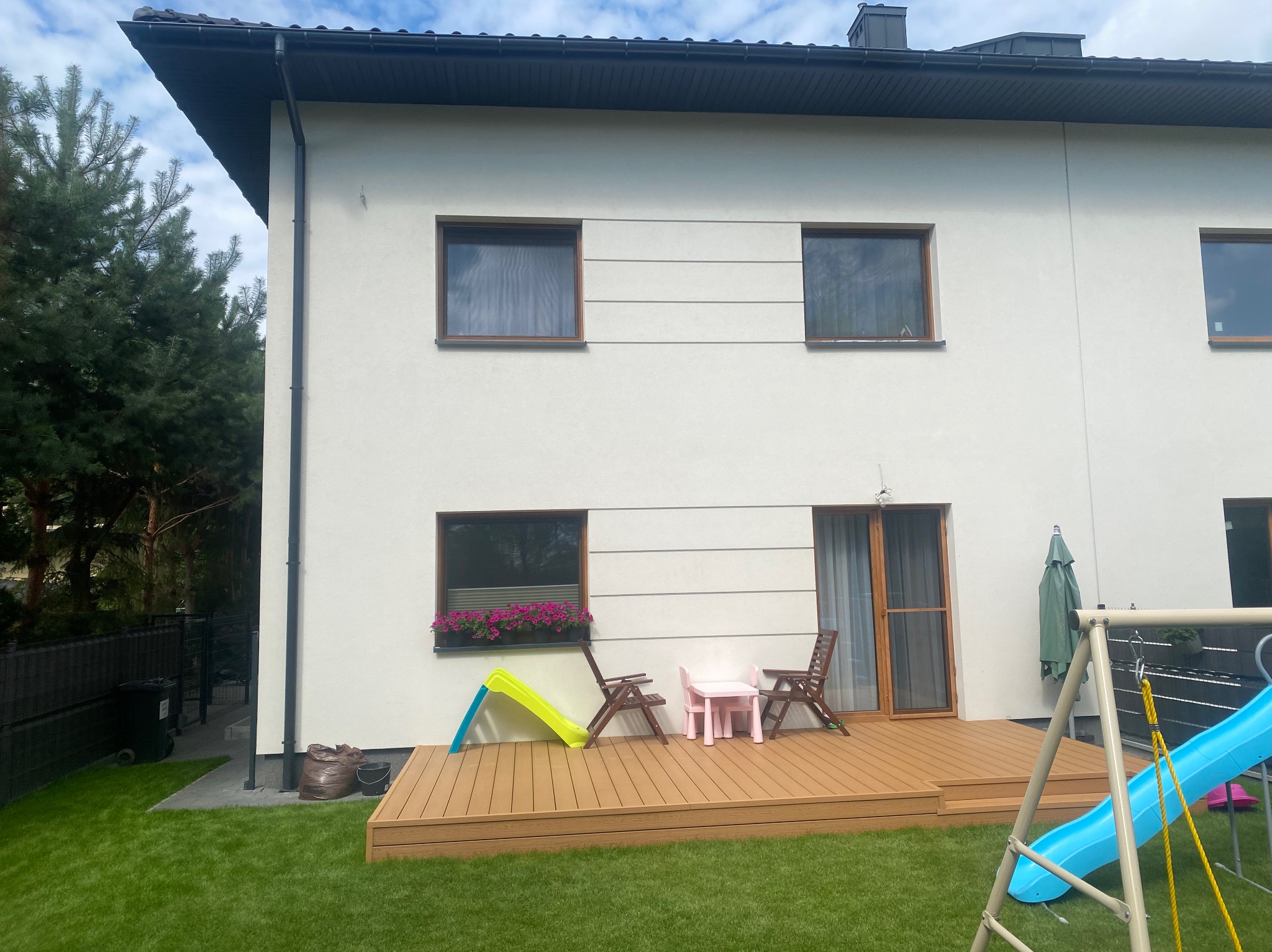 Dwupiętrowy dom z tarasem z desek kompozytowych, dziecięcym stolikiem i zjeżdżalnią. Widok na trawnik i roślinność. Nowoczesne wykończenie elewacji.