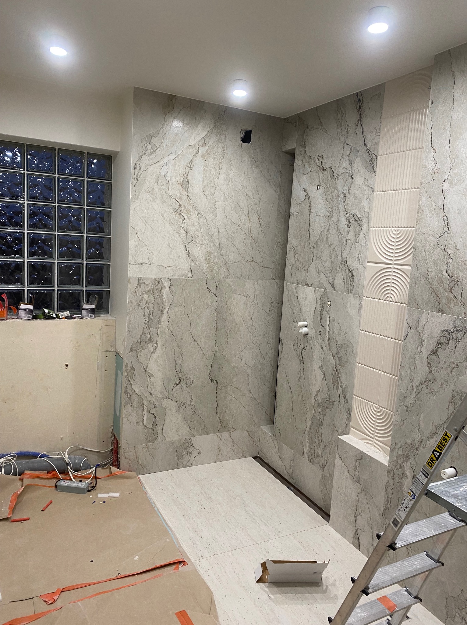 Wnętrze łazienki w trakcie remontu: marmurowe płytki ścienne, dekoracyjna listwa z motywem półkola, okno z luksferów, drabina i zabezpieczona podłoga.