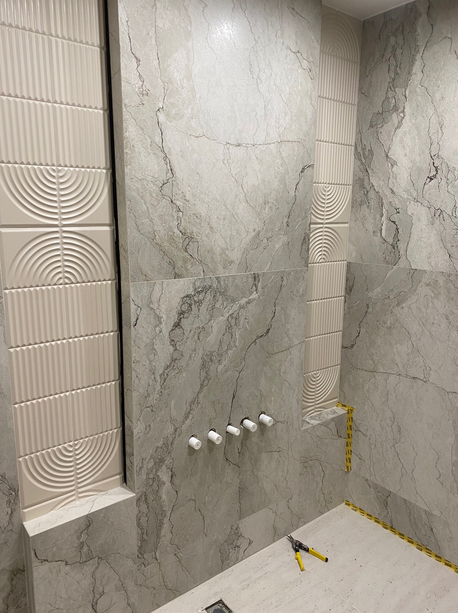 Wykończona ściana w łazience z dekoracyjnymi, beżowymi płytkami. Na ścianie widoczne wyprowadzenia instalacji wodnej. Na podłodze leżą nożyce do cięcia blachy.