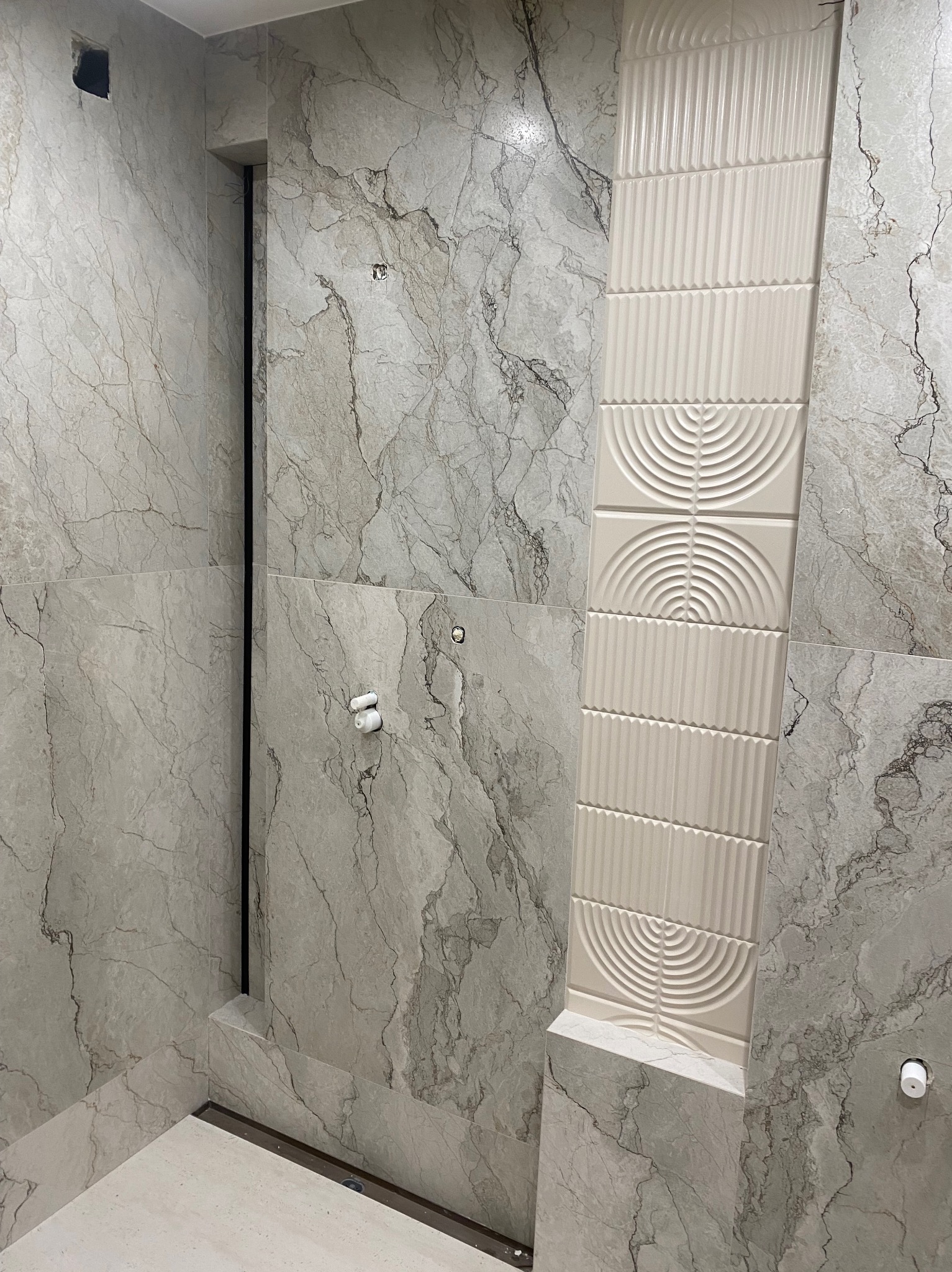 Wykończona łazienka z szarymi płytkami imitującymi marmur i dekoracyjnym pasem z białych, żłobionych płytek. Widoczne elementy instalacji i odpływ liniowy.