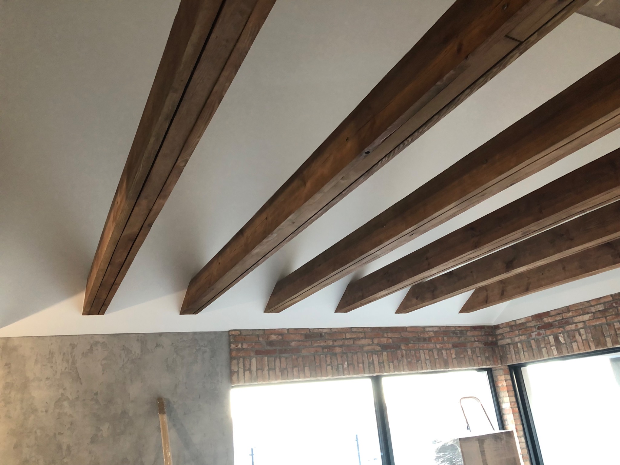 Jasne wnętrze z drewnianymi belkami stropowymi i fragmentem ściany z cegły. Widoczna ściana z szarą fakturą. Duże okno w tle.