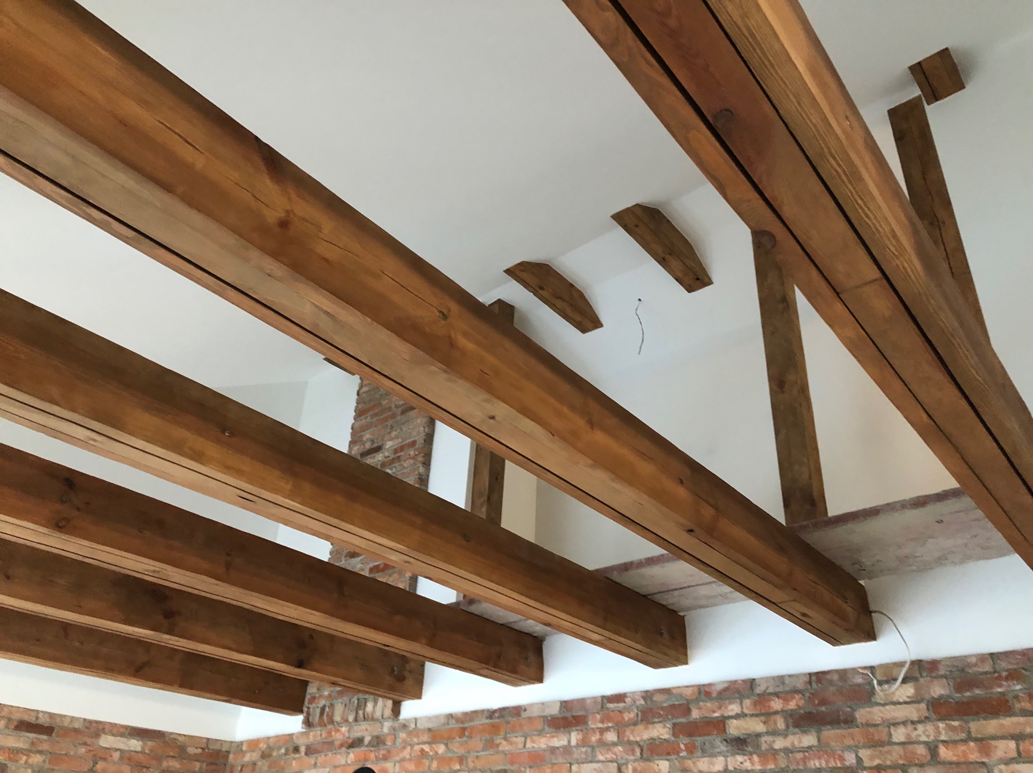 Drewniane belki stropowe, widoczne od dołu, w jasnym wnętrzu z fragmentem ściany z cegły. Konstrukcja w trakcie montażu, widoczne przewody.