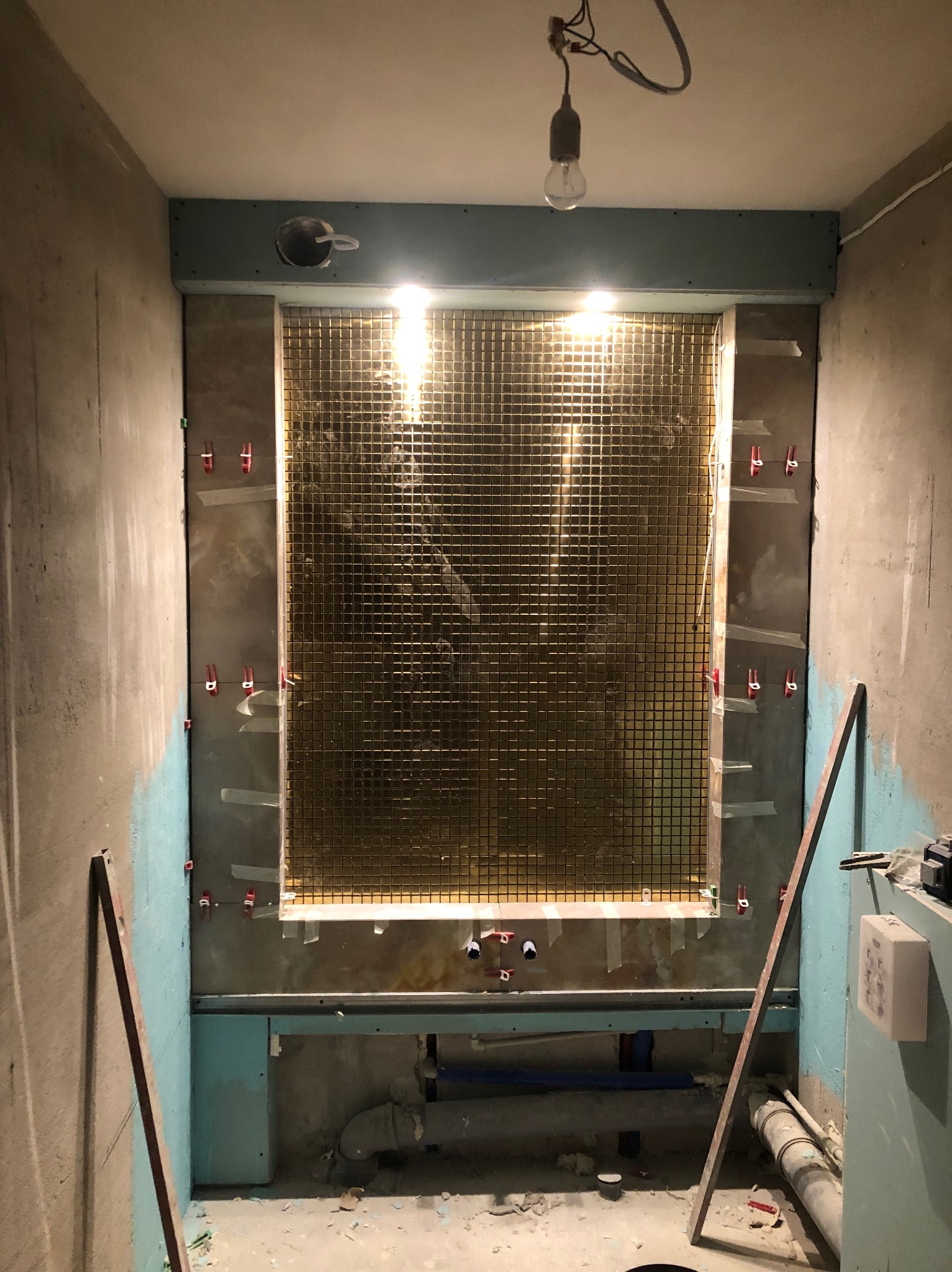 Łazienka w trakcie remontu: metalowa mozaika na ścianie, poziomica, rury instalacyjne. Widoczne elementy montażowe i surowy stan wykończenia wnętrza.
