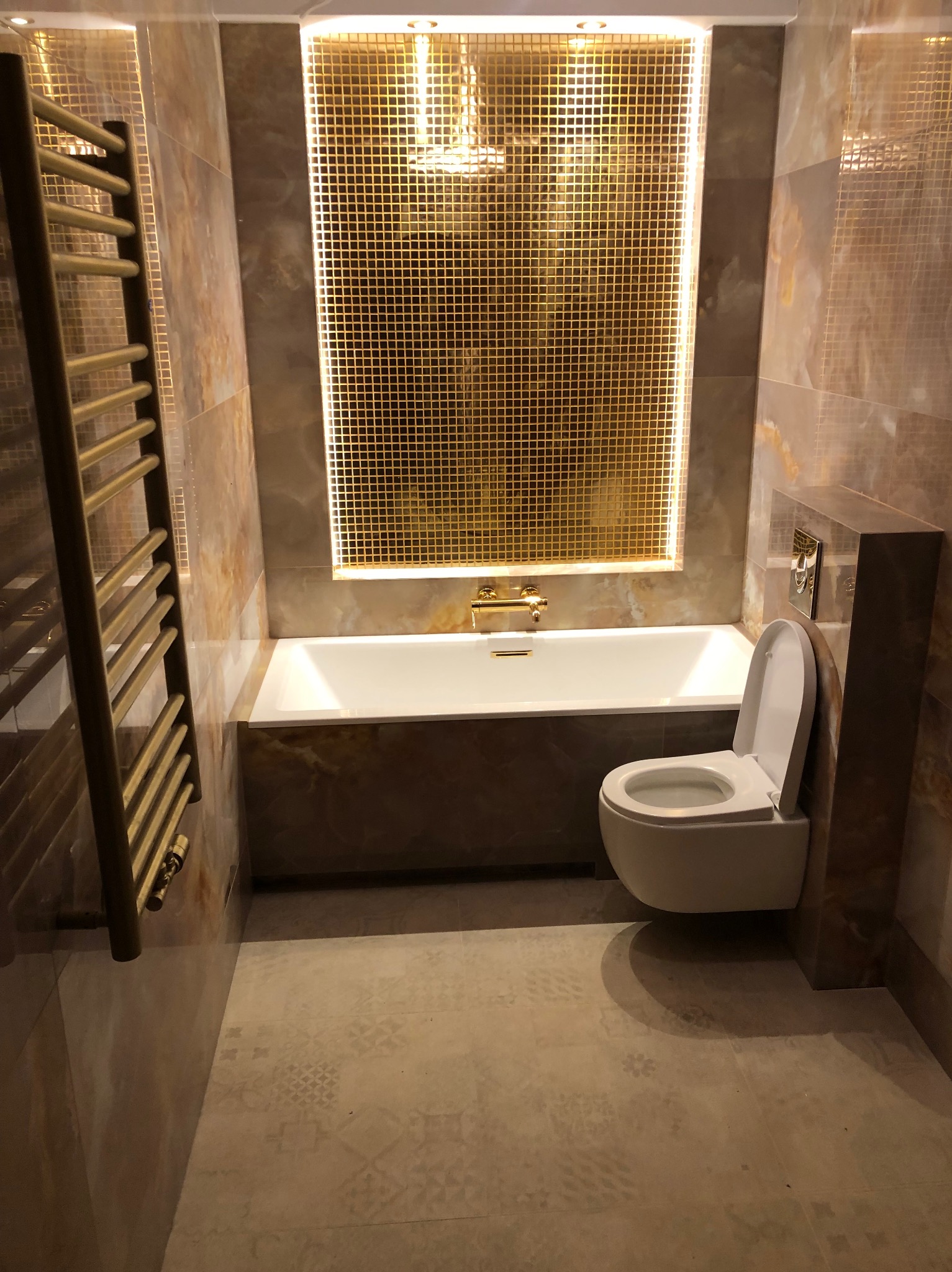 Luksusowa łazienka w odcieniach beżu i złota. Umywalka, toaleta i grzejnik w złotym kolorze. Ściany i podłoga wykończone płytkami imitującymi marmur.