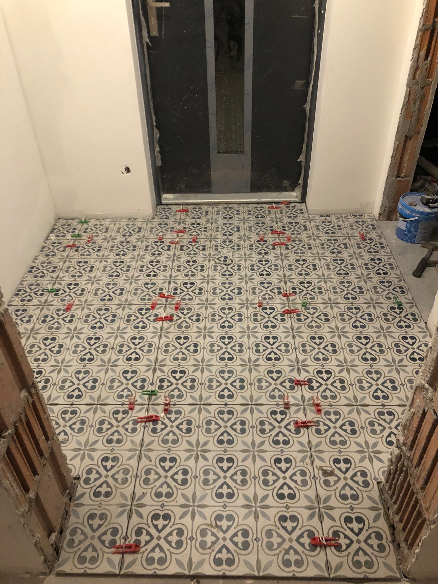 Układanie wzorzystych płytek podłogowych z użyciem poziomujących klipsów i klinów w przedpokoju, widoczne surowe wykończenia ścian i futryny drzwi.