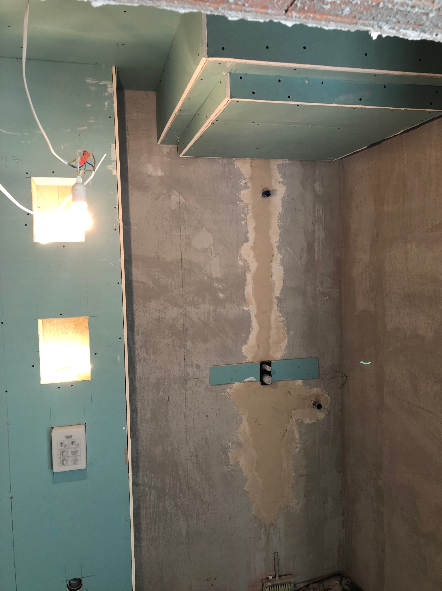 Łazienka w trakcie remontu: ściany z płyt karton-gips, odsłonięta instalacja elektryczna, sufit podwieszany z widoczną konstrukcją. Oświetlenie tymczasowe.
