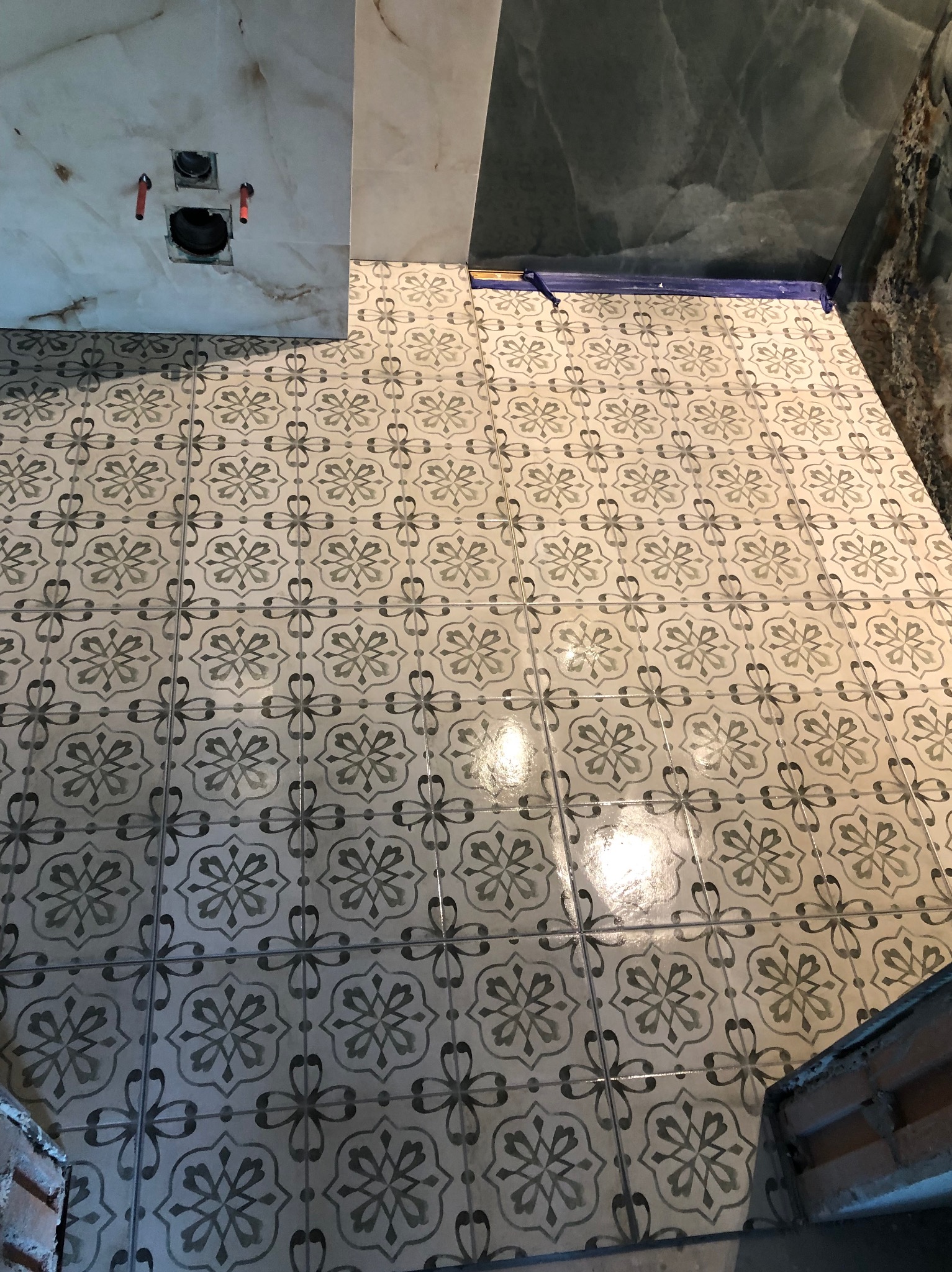 Ukończona podłoga w łazience z wzorzystymi płytkami w odcieniach szarości i beżu, fragment ściany wyłożonej płytkami imitującymi marmur oraz przygotowane podejście pod WC.