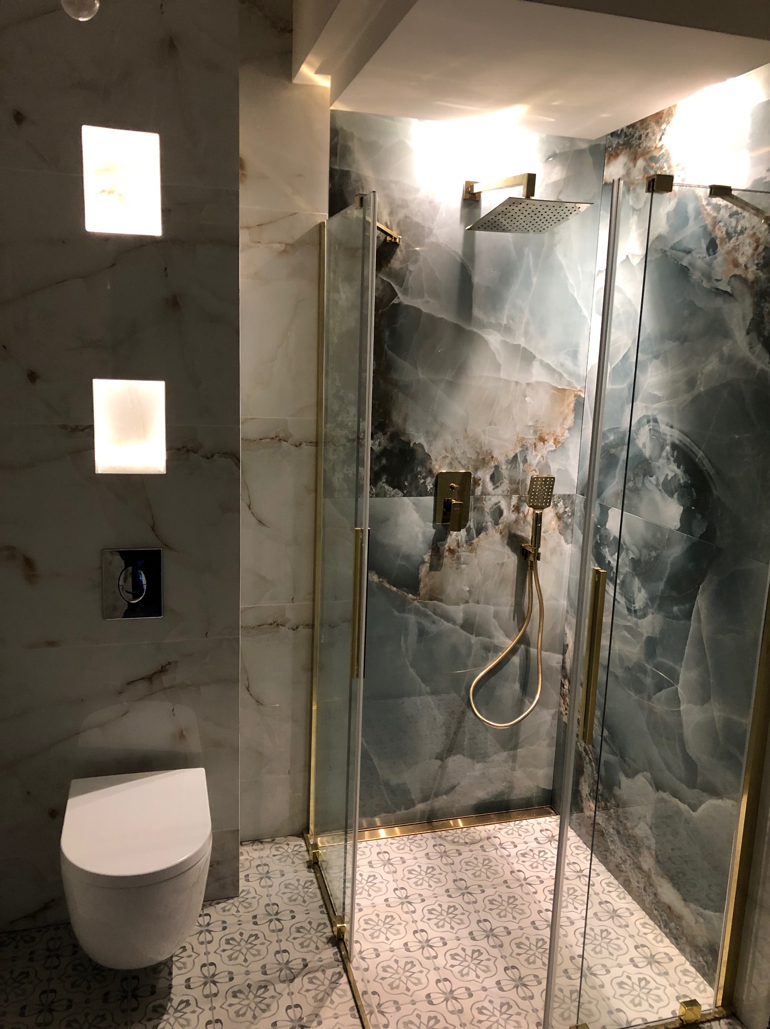 Nowoczesna łazienka z kabiną prysznicową typu walk-in, marmurowe płytki, złote detale i podświetlane lustra. Uwagę przykuwają wzorzyste płytki podłogowe.