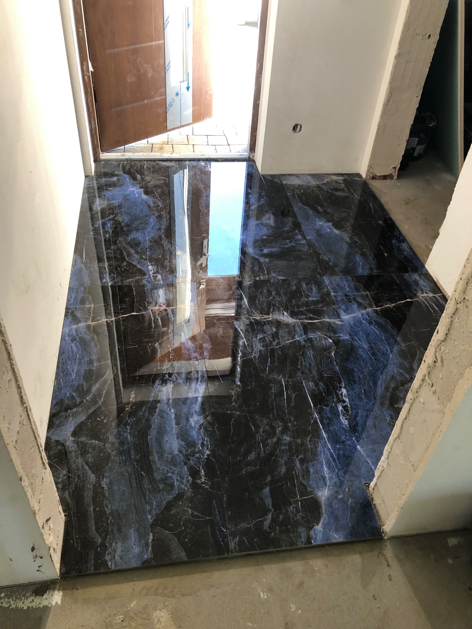 Połyskująca, nowa podłoga z ciemnych płytek imitujących marmur, ułożona w korytarzu. Widoczny fragment otwartych drzwi i surowy stan wykończenia ścian.