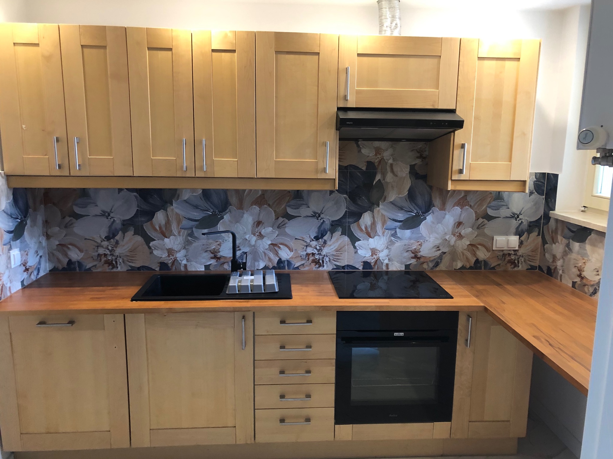 Jasna kuchnia z drewnianymi frontami, czarnym zlewem i płytą. Nad blatem dekoracyjne płytki z motywem kwiatowym. Nowoczesny design.